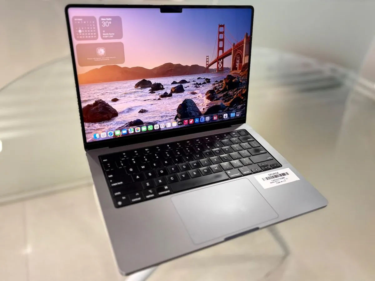 MacBook Pro 14-inch M1 Pro in Silver from a front-left three-quarter angle, open with the display on and the SPURGE asset label on the palm rest